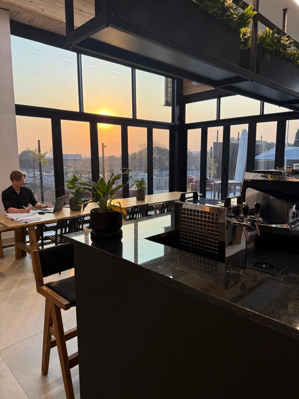 Café area at the Seventh Star venue with a granite counter and bar seating, looking out through full-height windows to a sunset over the treeline