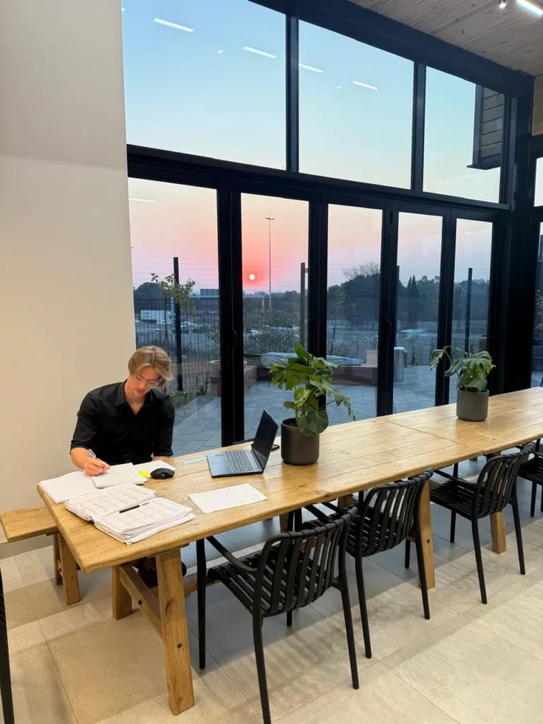 Long timber communal table beside full-height windows at the Seventh Star venue, with bench and chair seating and a person working at a laptop