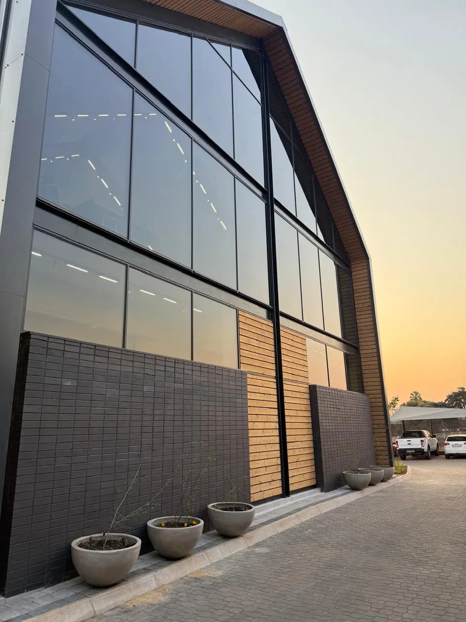 Double-volume glass facade of the Seventh Star venue at sunset, with grey brick and timber cladding and concrete planters along the paved walkway