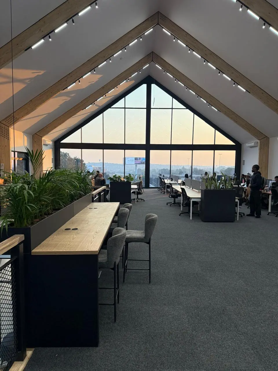 Open-plan mezzanine workspace at the Seventh Star venue with a high bar-height table and stools, planters dividing the desks, and a pitched gable window at the far end